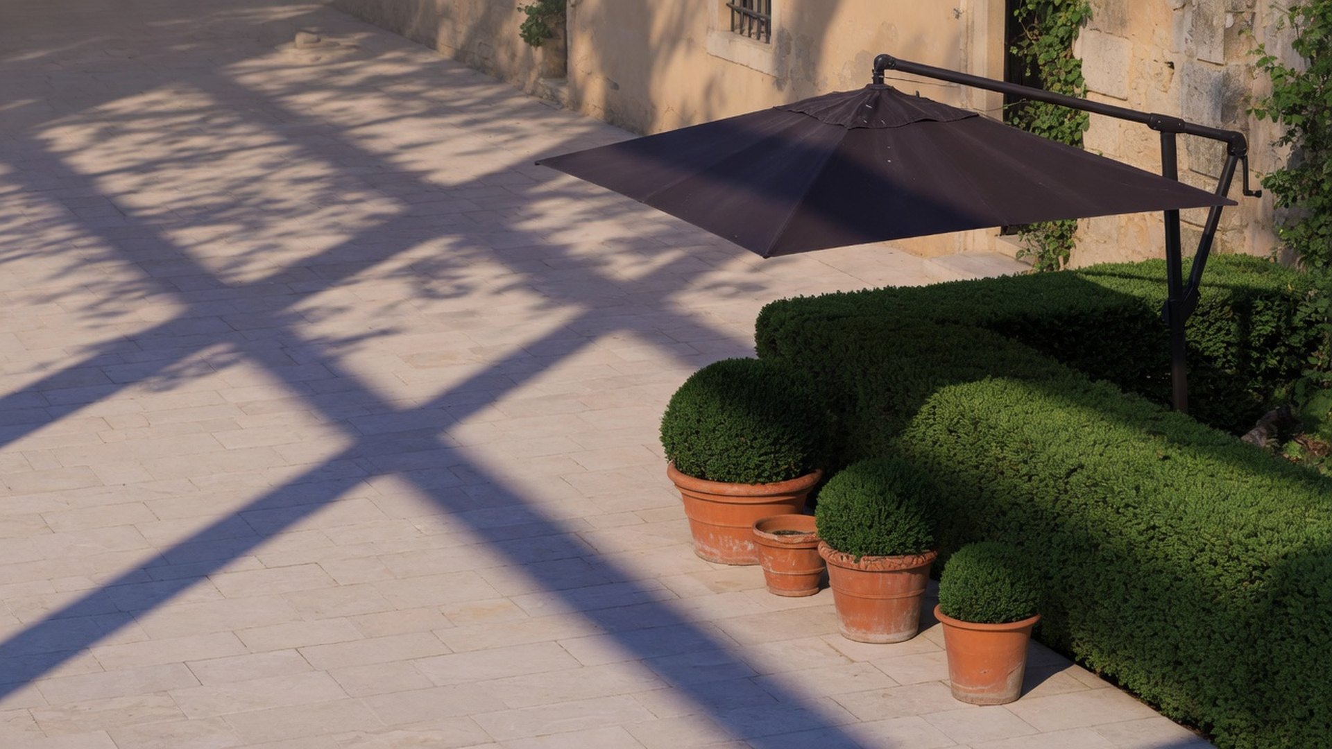 Late-afternoon limestone courtyard with long tree shadows, clipped hedges, terracotta pots and a dark cantilever parasol.